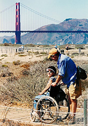 An able-bodied man walks with a woman in a wheelchair with the Golden Gate Bridge in the background
