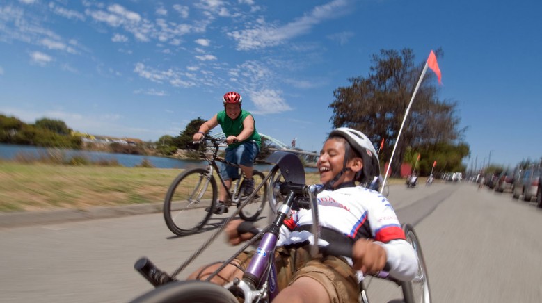 A young adaptive cyclist enjoys a ride with a friend on a conventional bicycle under blue skies