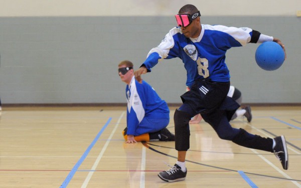 A goalball player about to throw the ball down the floor with a teammate in the background
