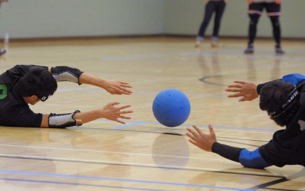 A goalball rolling right between the outstretched hands of two defenders