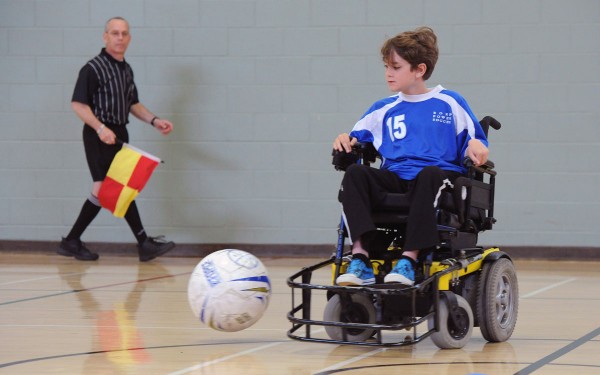 A BORP youth power soccer player dribbles a ball while a ref looks on