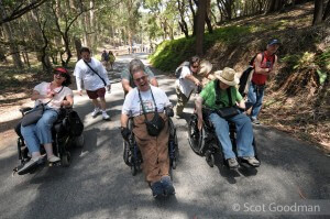 Three people in wheelchairs with able-bodied volunteers behind share a laugh on a shady