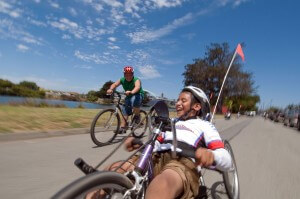 A young adaptive cyclist enjoys a ride with a friend on a conventional bicycle under blue skies