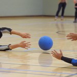 A goalball rolling right between the outstretched hands of two defenders