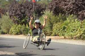 A woman riding an adaptive cycle raises her hands triumphantly