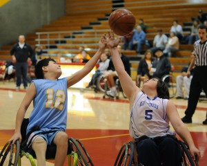 Two youth wheelchair basketball players reach for a basketball in the air above them