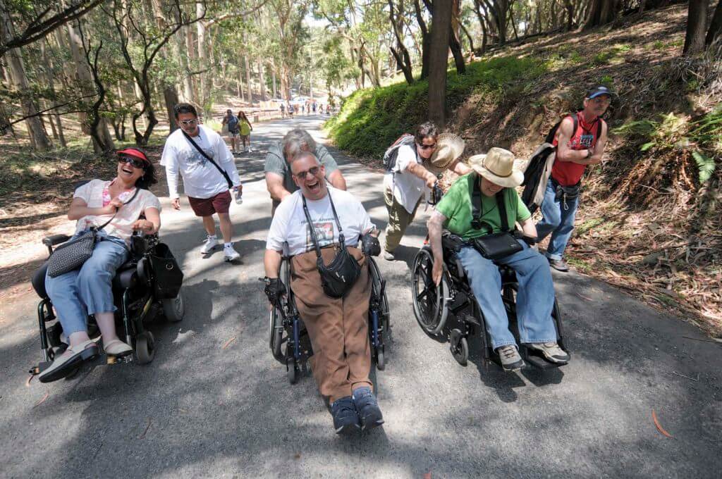 Three people in wheelchairs with volunteers standing behind them laughing on a hike.