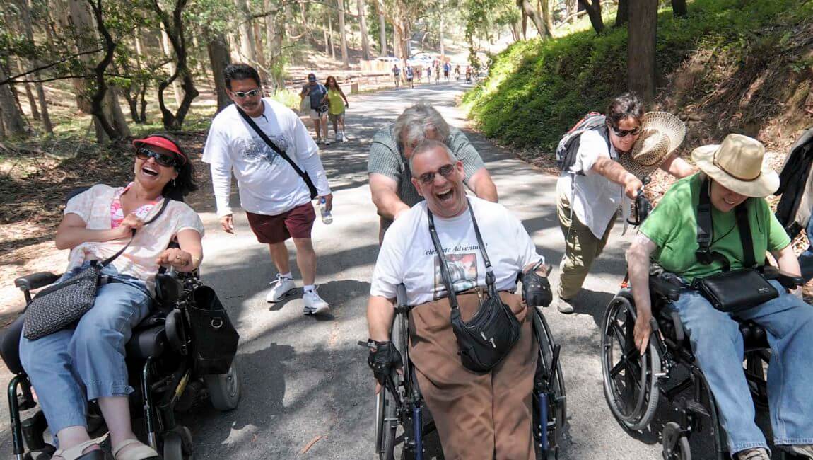 Three people in wheelchairs with volunteers standing behind them laughing on a hike.
