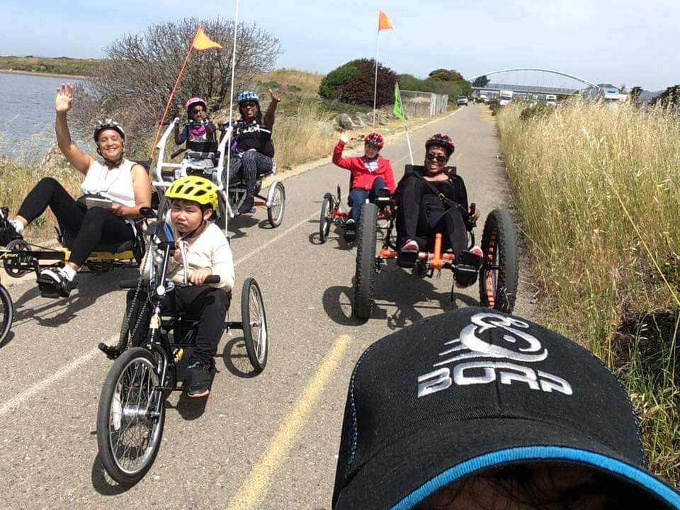 Group of smiling adults and children on adaptive bikes ride trail together