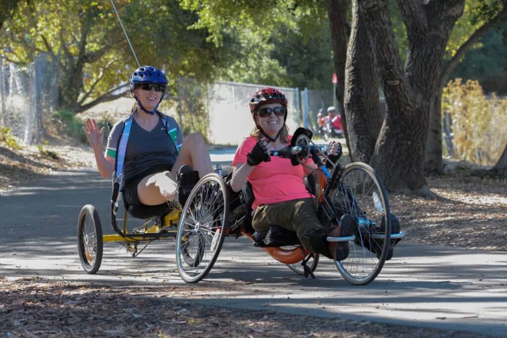 Handcyclist and support rider smiling and waving as they ride on trail