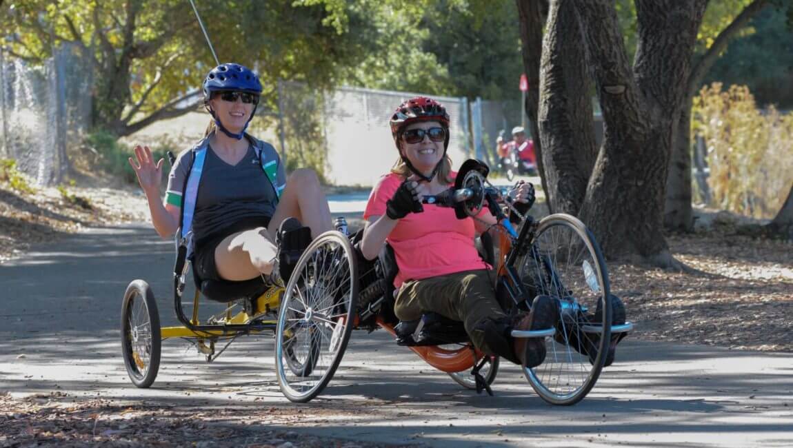 Handcyclist and support rider smiling and waving as they ride on trail