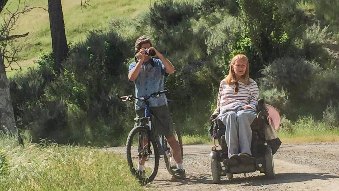 One person in a wheelchair and one person with bike holding a camera on an accessible trail.