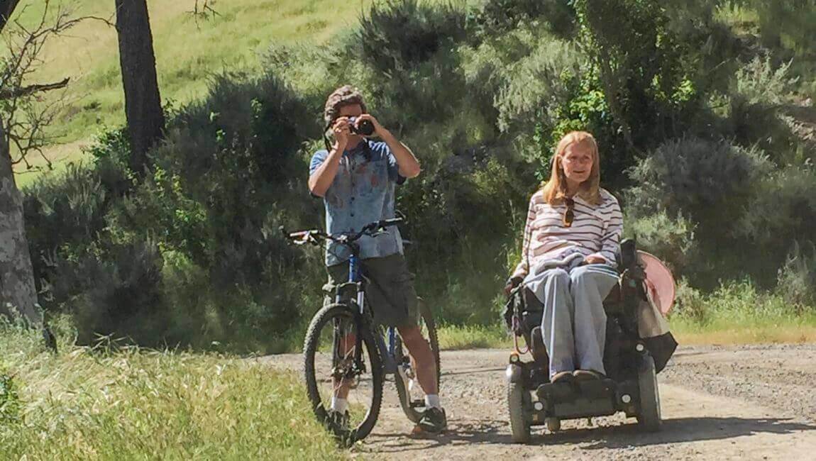 Una persona en silla de ruedas y una persona en bicicleta sosteniendo una cámara en un sendero accesible.