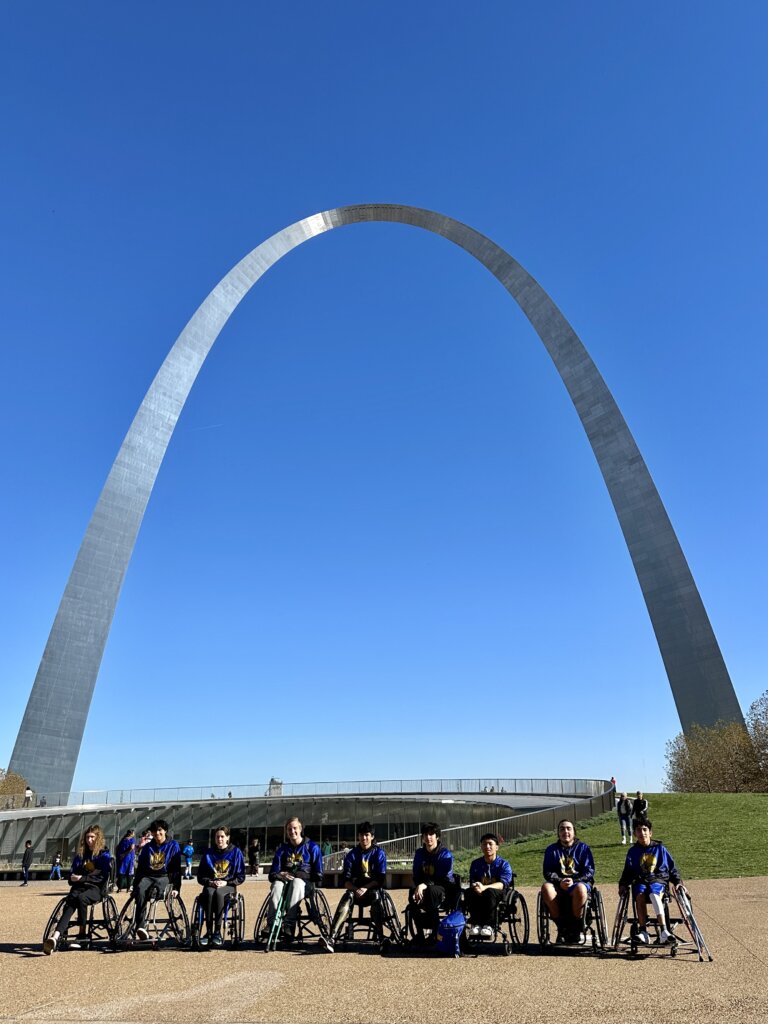 Nine Jr. Road Warriors posing in their wheelchairs in front of the St Louis Arch, which makes a silver arch against a blue blue sky.