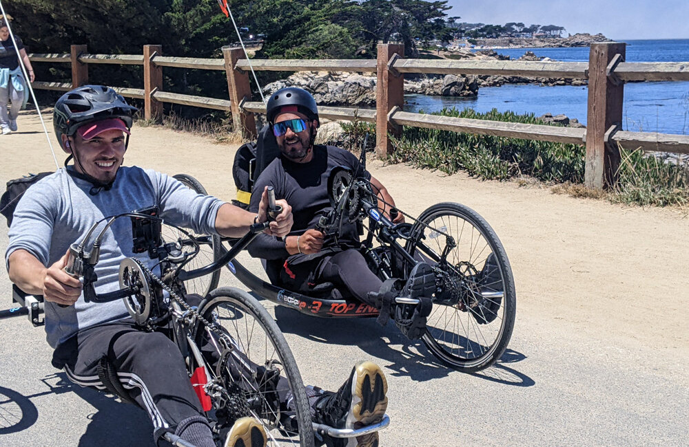 two young men on hand cycles are smiling while riding by a fence. the ocean is behind them