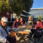 A group of people of different ages and ethnicities in wheelchairs gathered around a table and looking at the camera.