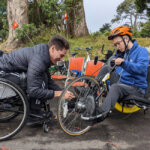 one person in a wheelchair is adjusting the foot pedals for another person who in an a hand tricycle