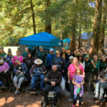Image1: 23 people gathered at a campsite, five are in wheelchairs and at least 6 are wearing green East Bay Parks t-shirts.