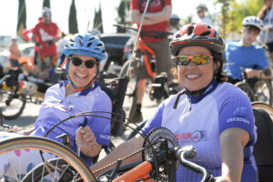 Two women, one in a recumbent cycle and one on a hand-cycle, both wearing the 2016 Revolution Ride.