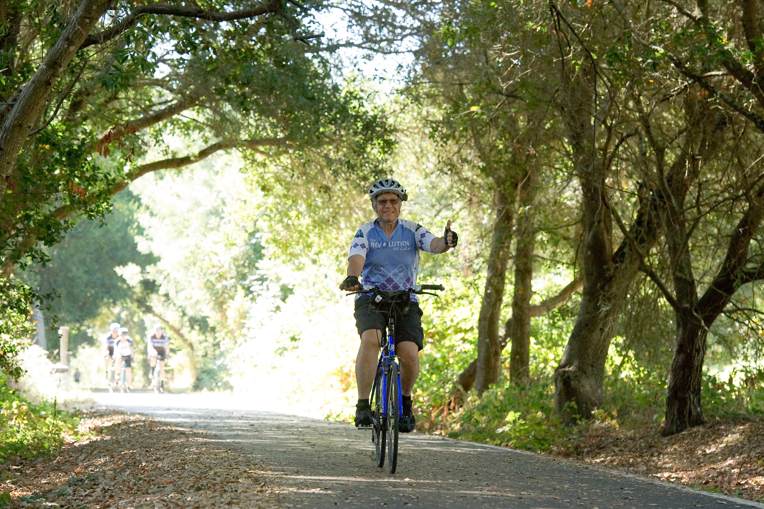 Cyclist riding through the trees giving a thumbs up and wearing a 2019 Revolution Ride jersey.