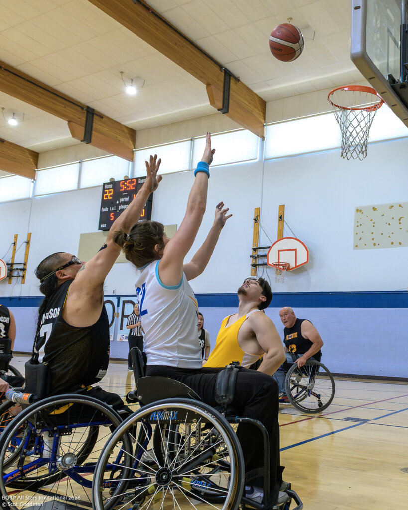 One wheelchair basketball player shoots with a defender behind her trying to block. The ball has left her hand and is on its way to the net