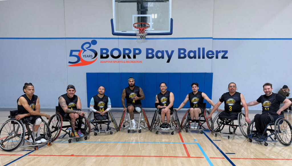 Eight basketball players using wheelchairs in black jerseys pose in front of a basketball hoop. Text above them reads BORP Bay Ballerz