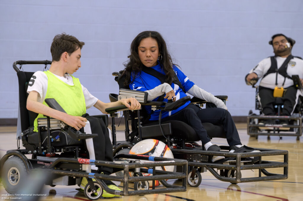 Two people on opposing teams in power soccer wheelchairs move parallel to each other with a power soccer ball between them. They both have looks of concentration.