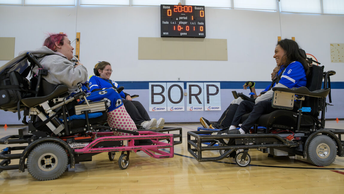 4 players in power soccer wheelchairs with guards on the front face each other smiling. The background shows 20 minutes on the game clock and a score of 0 to 0.