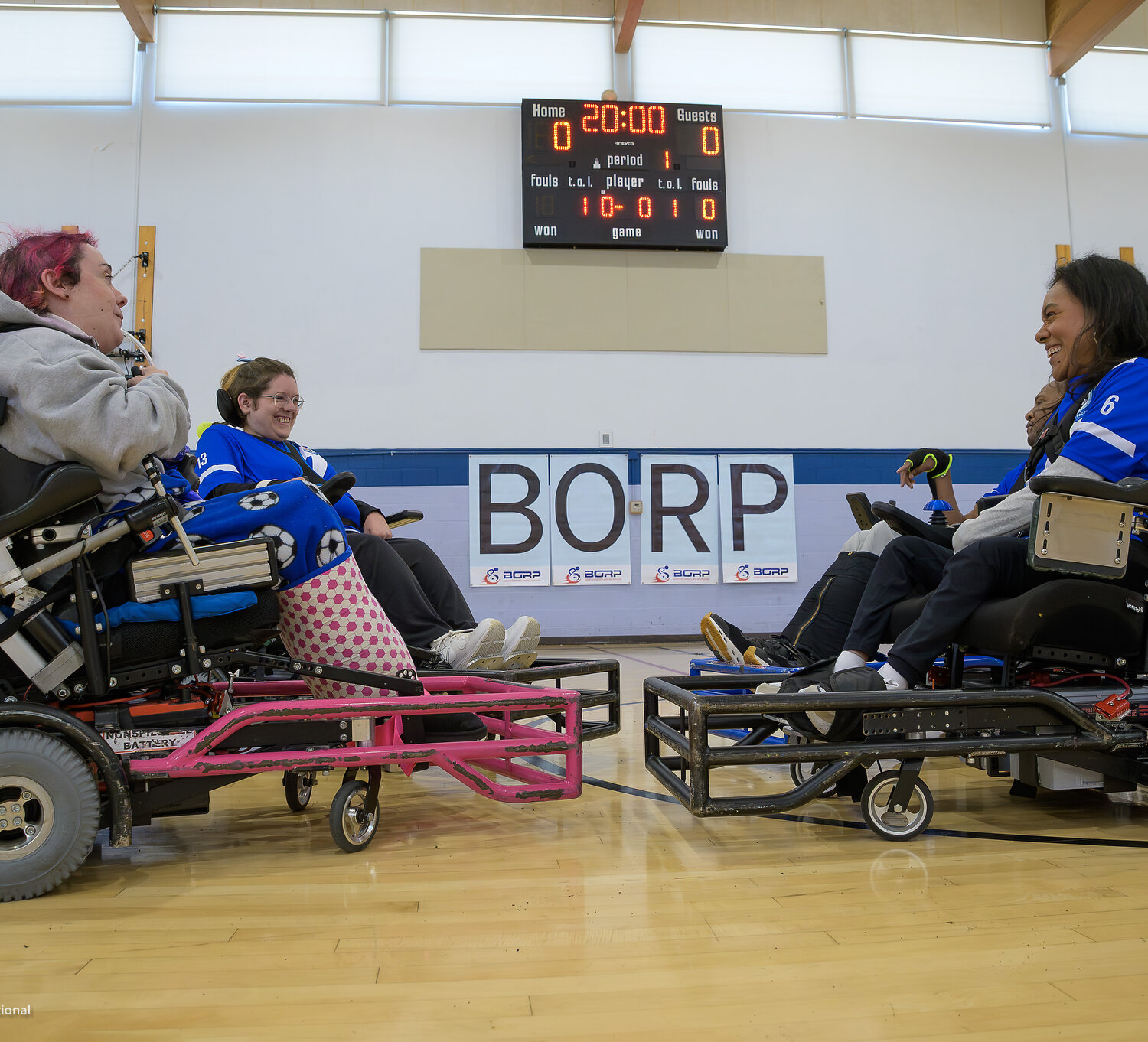 4 players in power soccer wheelchairs with guards on the front face each other smiling. The background shows 20 minutes on the game clock and a score of 0 to 0.