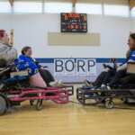 4 players in power soccer wheelchairs with guards on the front face each other smiling. The background shows 20 minutes on the game clock and a score of 0 to 0.