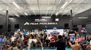 A large group of people pose in front of a sign reading Peak Pickleball. About half of the people use wheelchairs, and half are standing behind them.