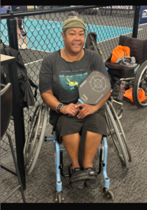 Woman poses for photo. She is sitting on a specialized pickleball wheelchair and holding a paddle.