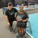 Two people holding pickleball paddles pose for photo. One person kneels next to the other person, who is sitting on a specialized pickleball wheelchair