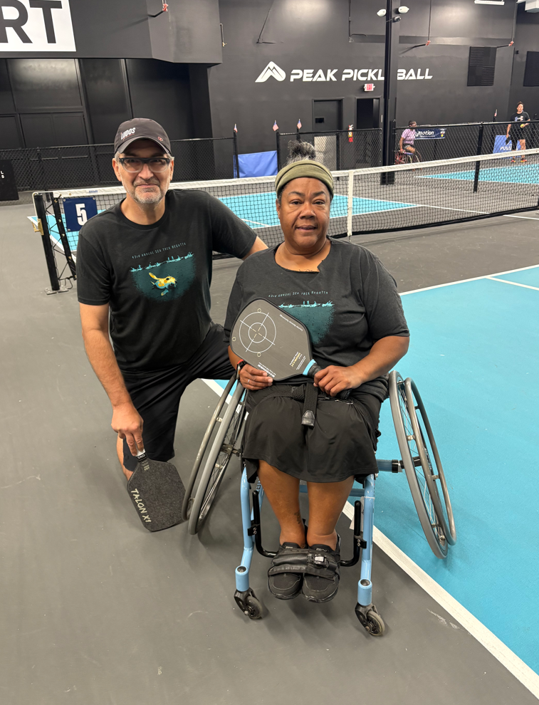 Two people holding pickleball paddles pose for photo. One person kneels next to the other person, who is sitting on a specialized pickleball wheelchair