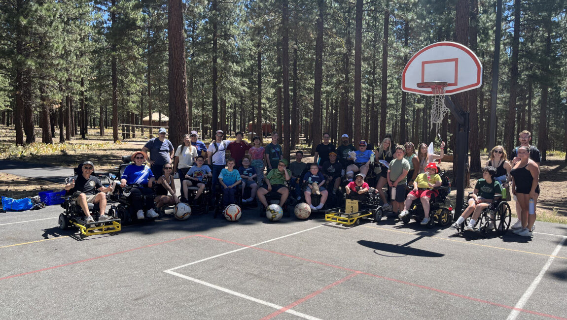 Power wheelchair users and camp ambulatory people posing on an outdoor basketball court with forest in the background
