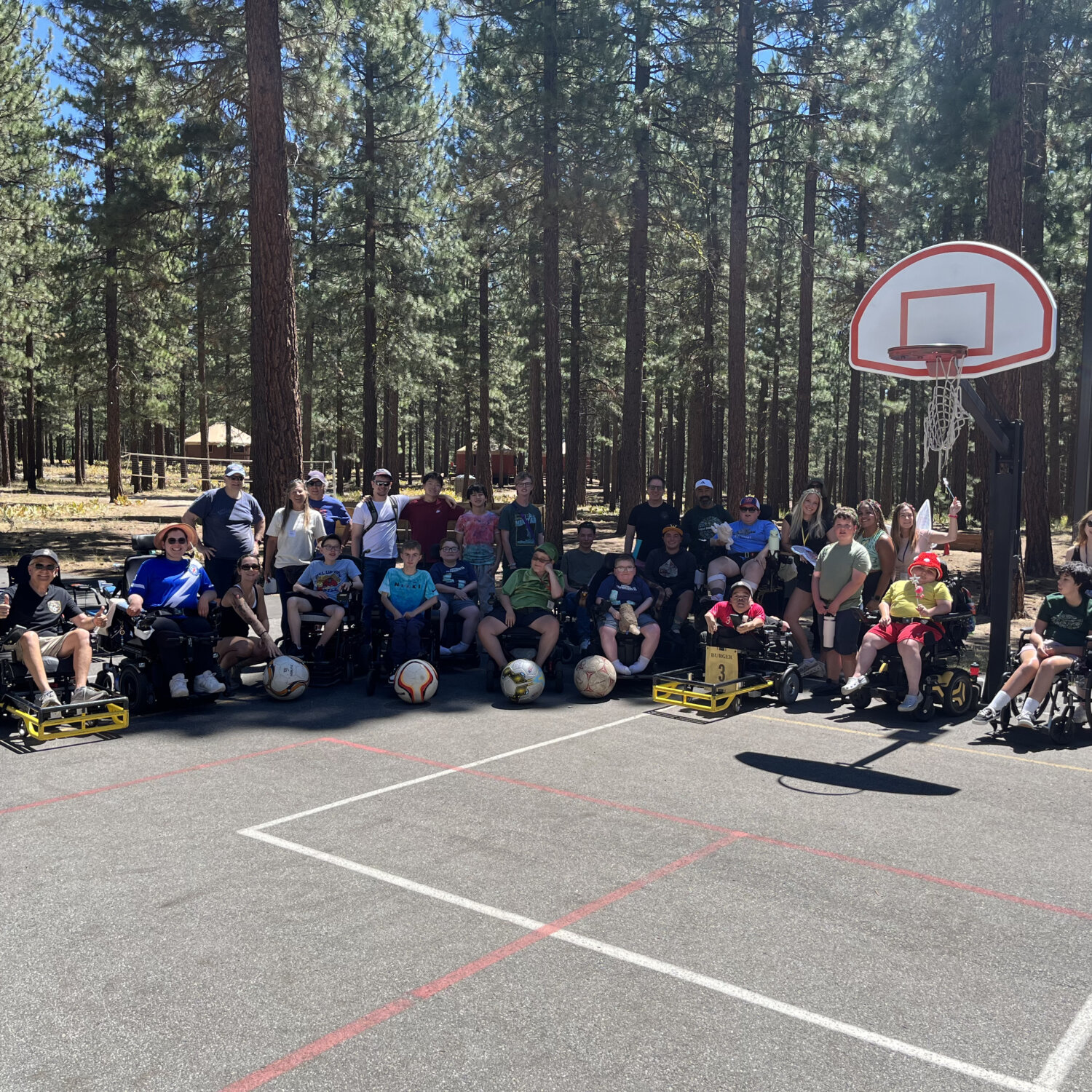 Power wheelchair users and camp ambulatory people posing on an outdoor basketball court with forest in the background