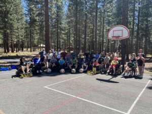 Power wheelchair users and camp ambulatory people posing on an outdoor basketball court with forest in the background